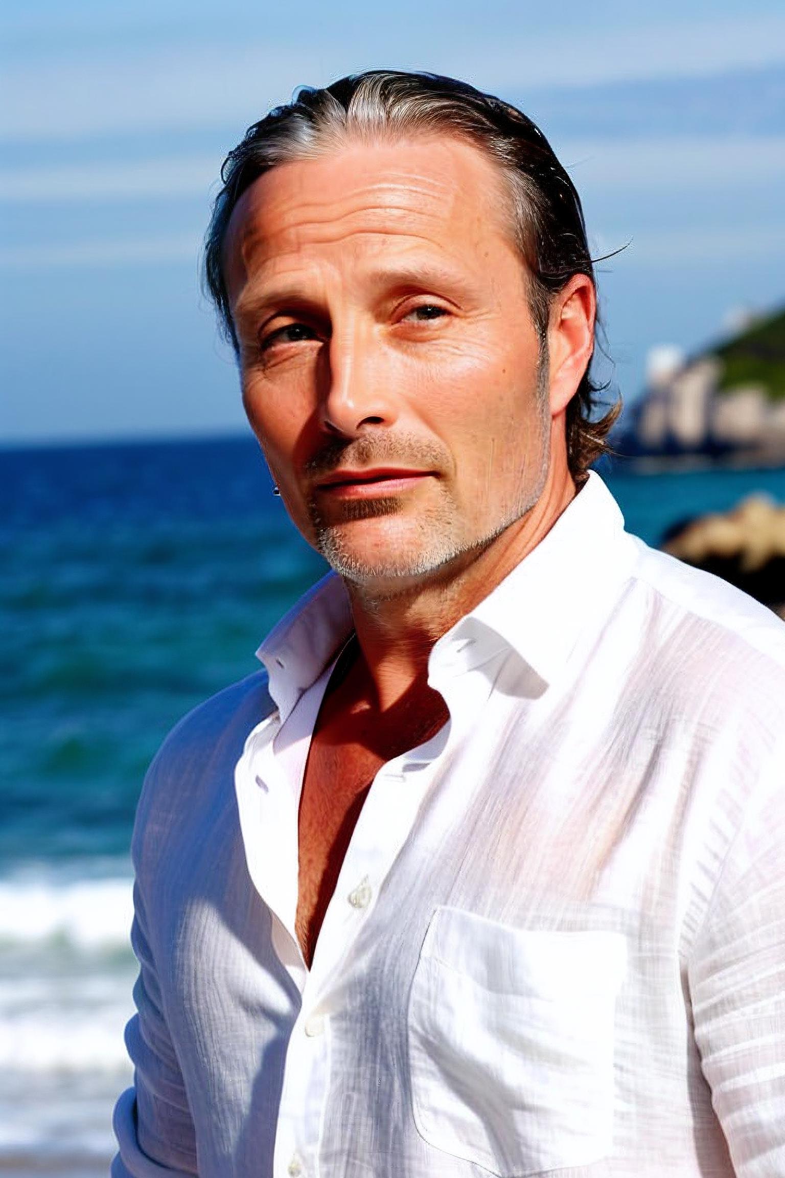 A man with a beard and wavy hair, wearing a white shirt and standing next to the ocean.